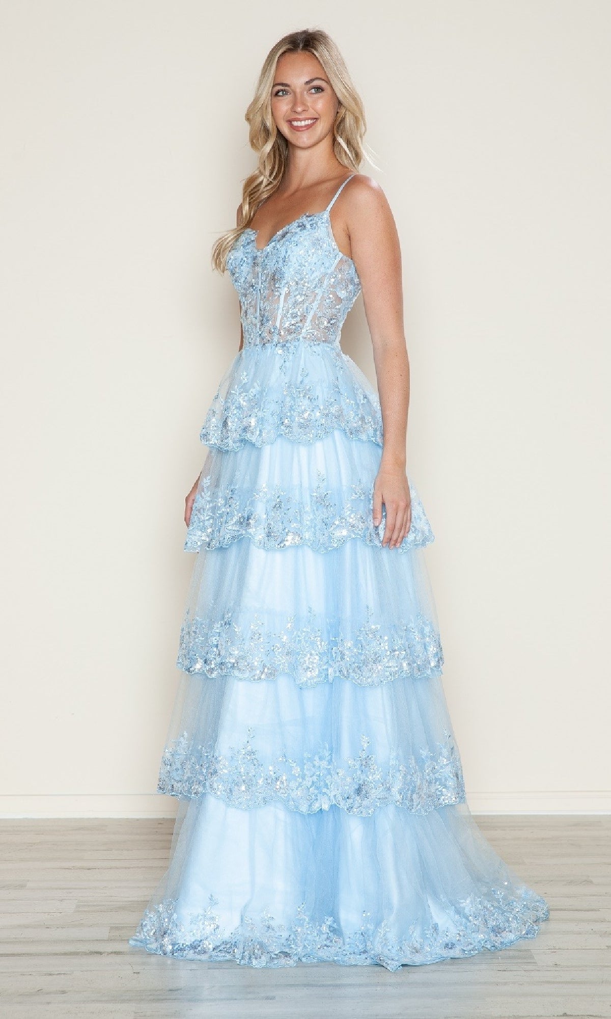 A woman smiles while wearing the blue Long Glitter-Lace Tiered Prom Dress (Poly USA 9548) featuring spaghetti straps and a semi-sheer bodice, standing against a plain light background.